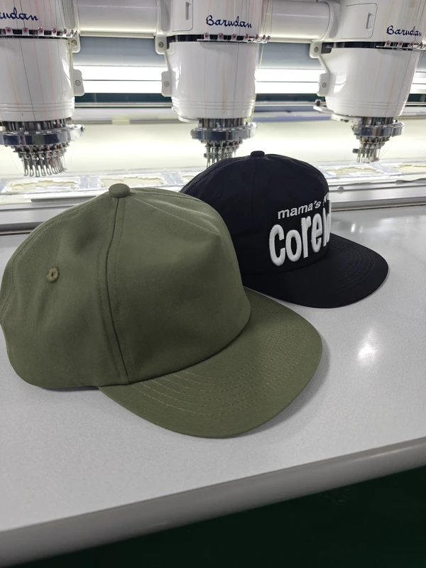 Olive green blank cap and black cap with white embroidery displayed at an embroidery workstation