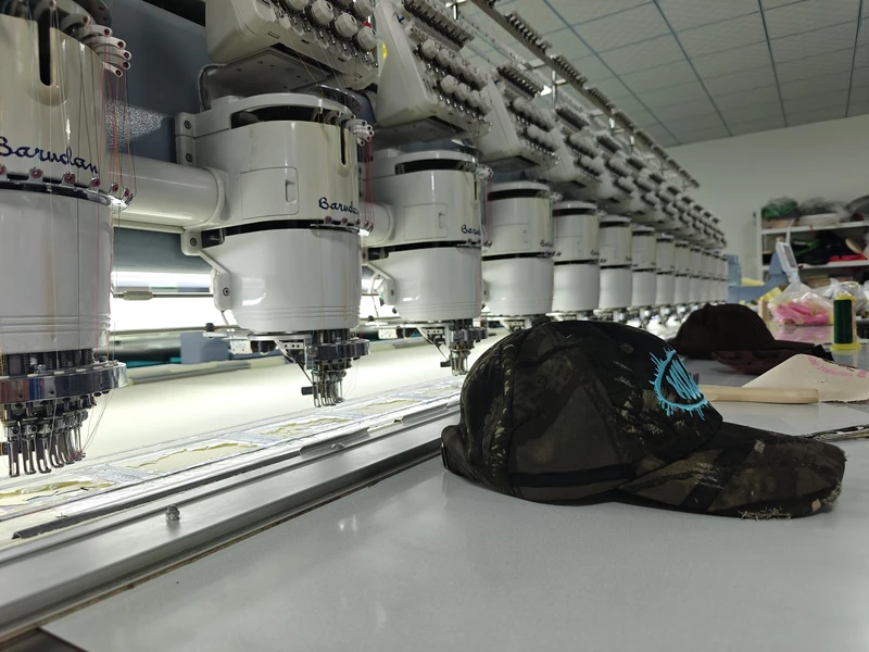 Camouflage cap with turquoise front embroidery resting beside multihead embroidery machines